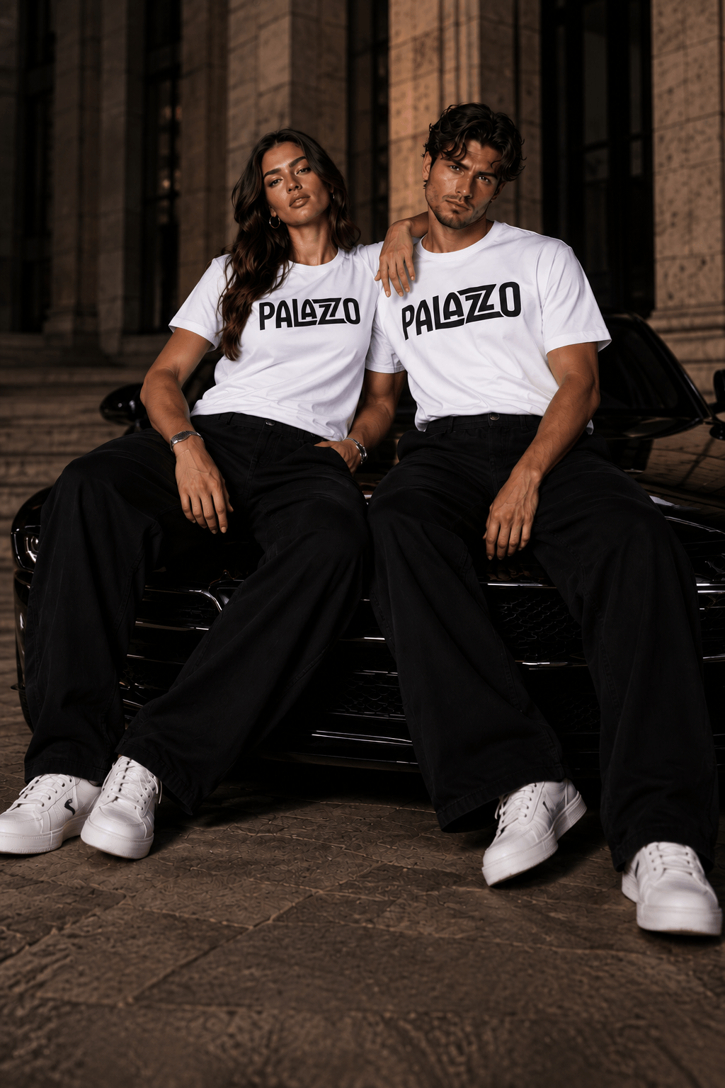 Zwei Models in weißen T-Shirts mit 'PALAZZO'-Schrift, sitzen auf einem schwarzen Auto vor einem historischen Gebäude, stilvolle Streetwear