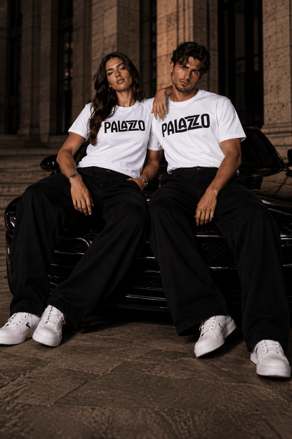 Zwei Models in weißen T-Shirts mit 'PALAZZO'-Schrift, sitzen auf einem schwarzen Auto vor einem historischen Gebäude, stilvolle Streetwear