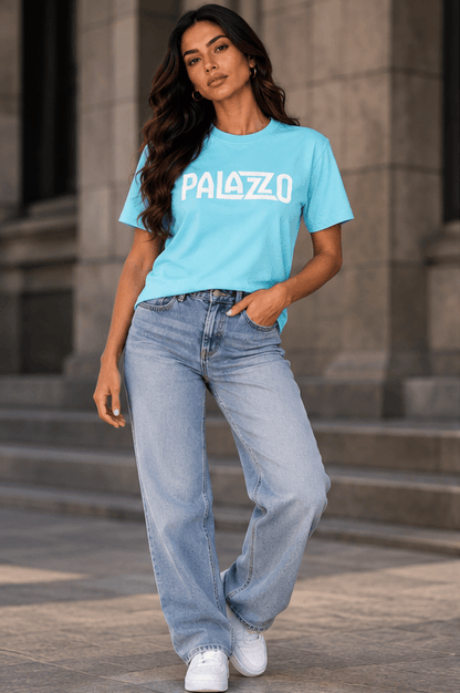 Hübsche Frau trägt ein hellblaues T-Shirt mit weißem Palazzo-Logo, lockere Jeans und weiße Sneakers vor einem urbanen Hintergrund